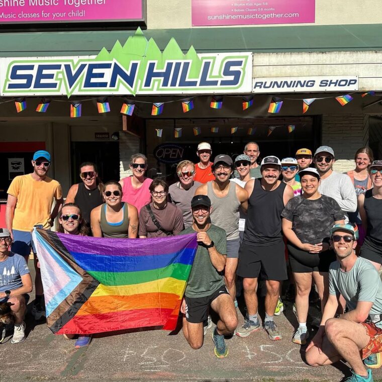 Out Trails group runners posing with a big rainbow Pride flag outside Seven Hills Running Shop.