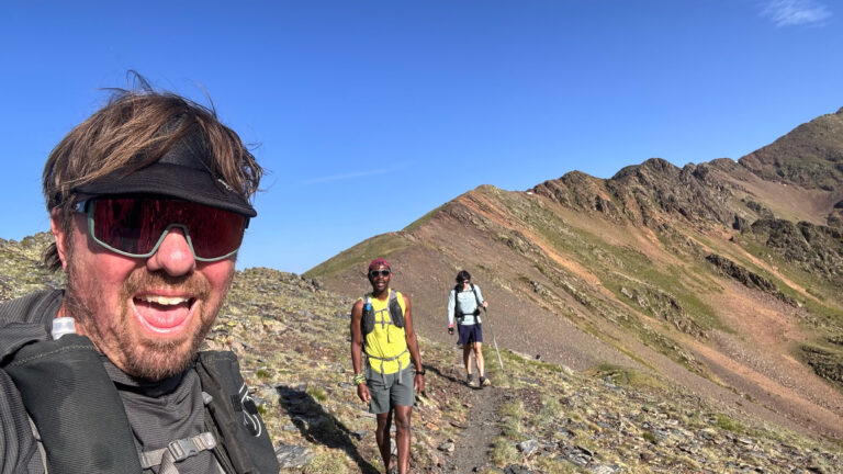 Ken Turner and two other runners on a rocky mountain trail in Iberia under a clear blue sky. Ken is smiling in the foreground wearing a visor and sunglasses. Two others in trail running gear are walking behind along a ridgeline.